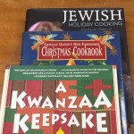 Three books stacked on top of each other, with cover titles visible: on bottom, Jewish Holiday Cooking; in the middle, Camille Glenn's Old Fashioned Christmas Cookbook; on top, A Kwanzaa Keepsake