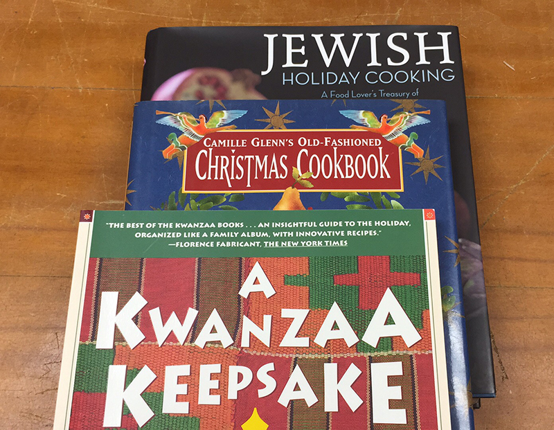 Three books stacked on top of each other, with cover titles visible: on bottom, Jewish Holiday Cooking; in the middle, Camille Glenn's Old Fashioned Christmas Cookbook; on top, A Kwanzaa Keepsake