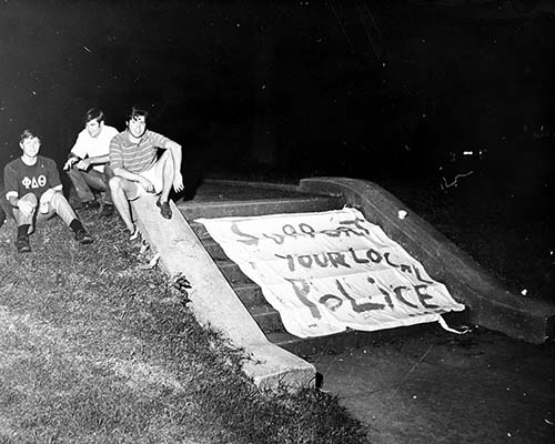 Male students sitting near a cloth sign draped over steps, reading Support Your Local Police